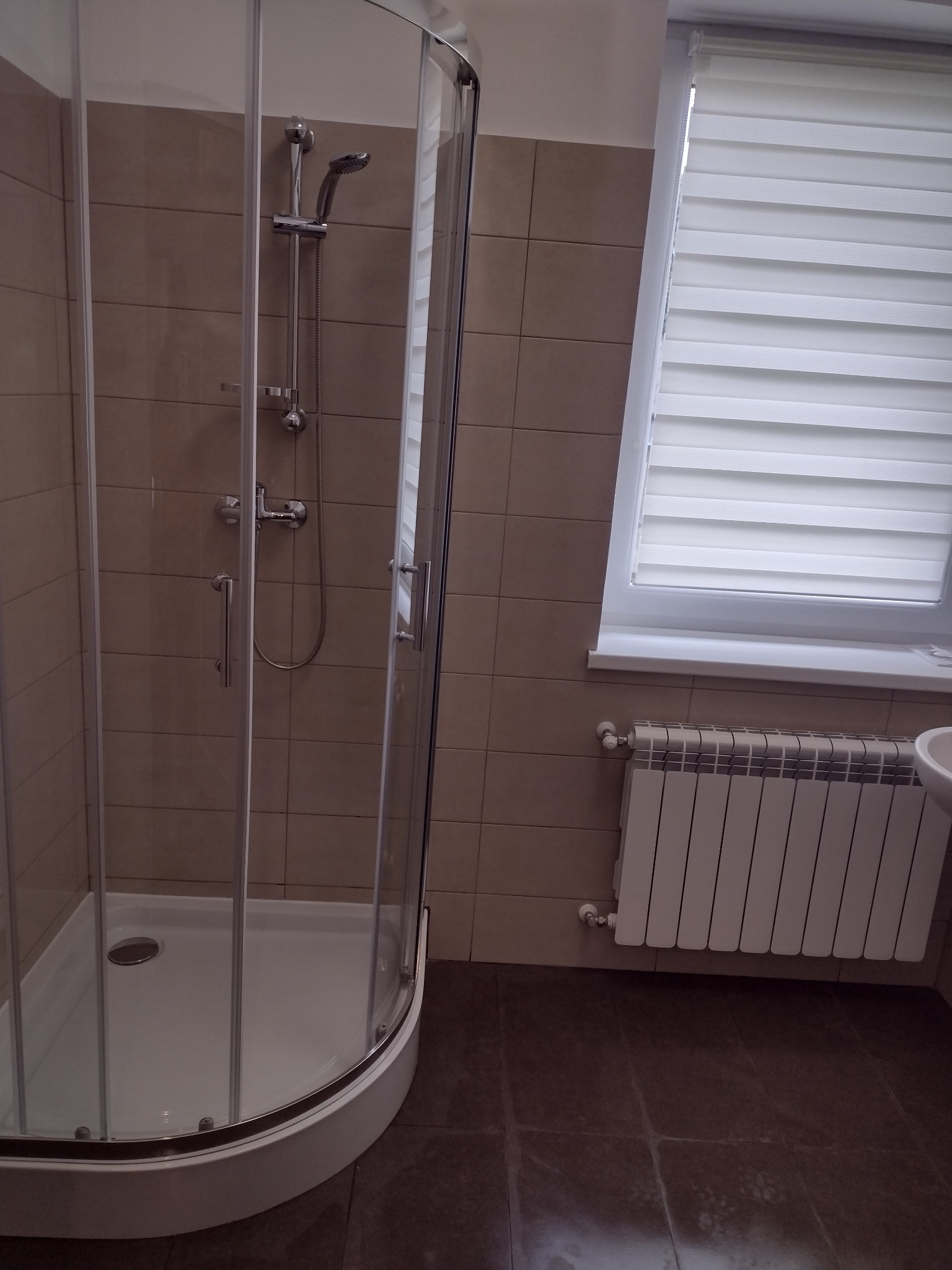 Narożna kabina prysznicowa z chromowaną armaturą na tle beżowych płytek, z widocznym oknem z żaluzjami i grzejnikiem.