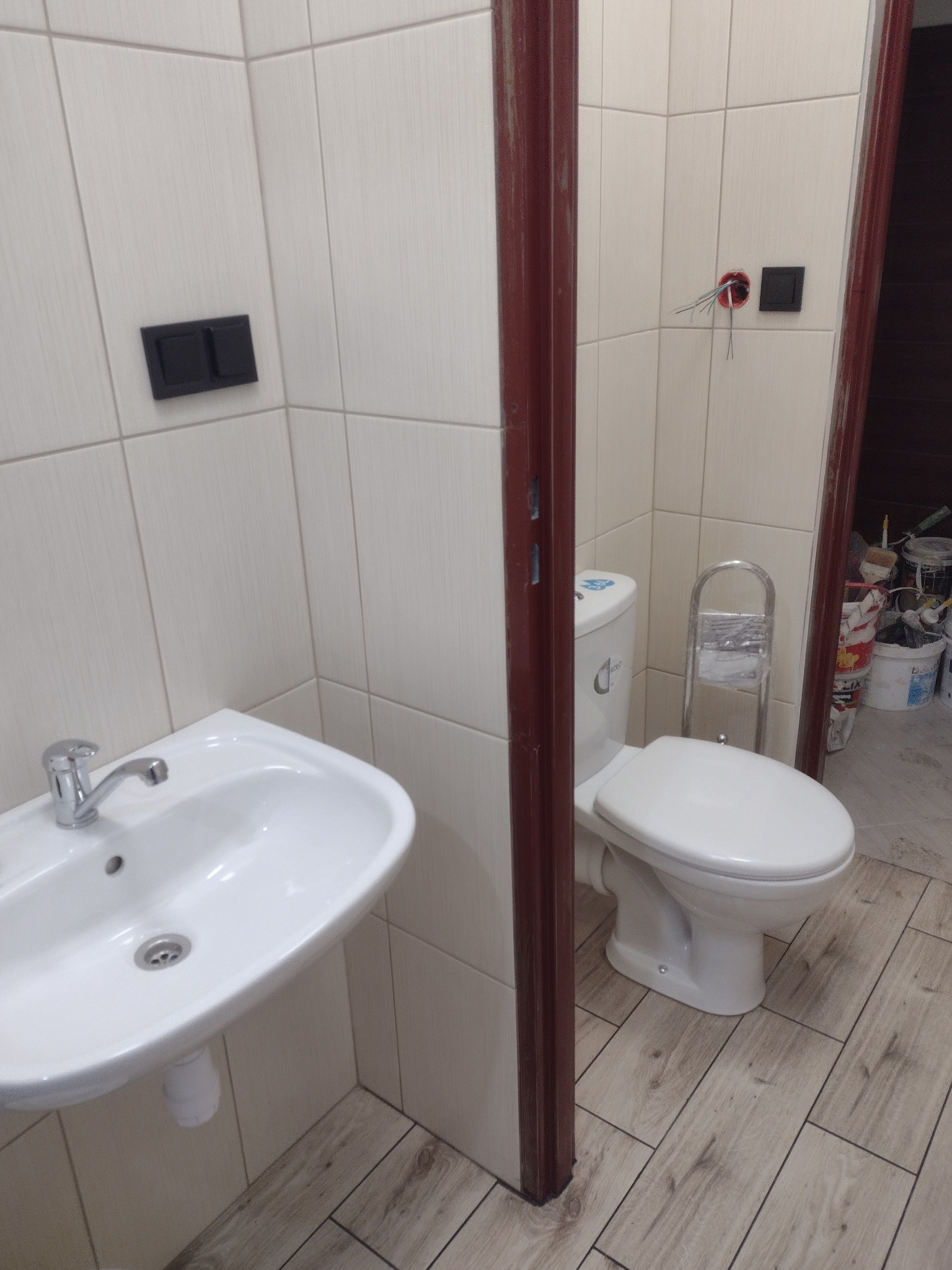 Wykończona łazienka z kremowymi płytkami, białą umywalką i toaletą, drewnopodobna podłoga, widoczne niedokończone gniazdko elektryczne.