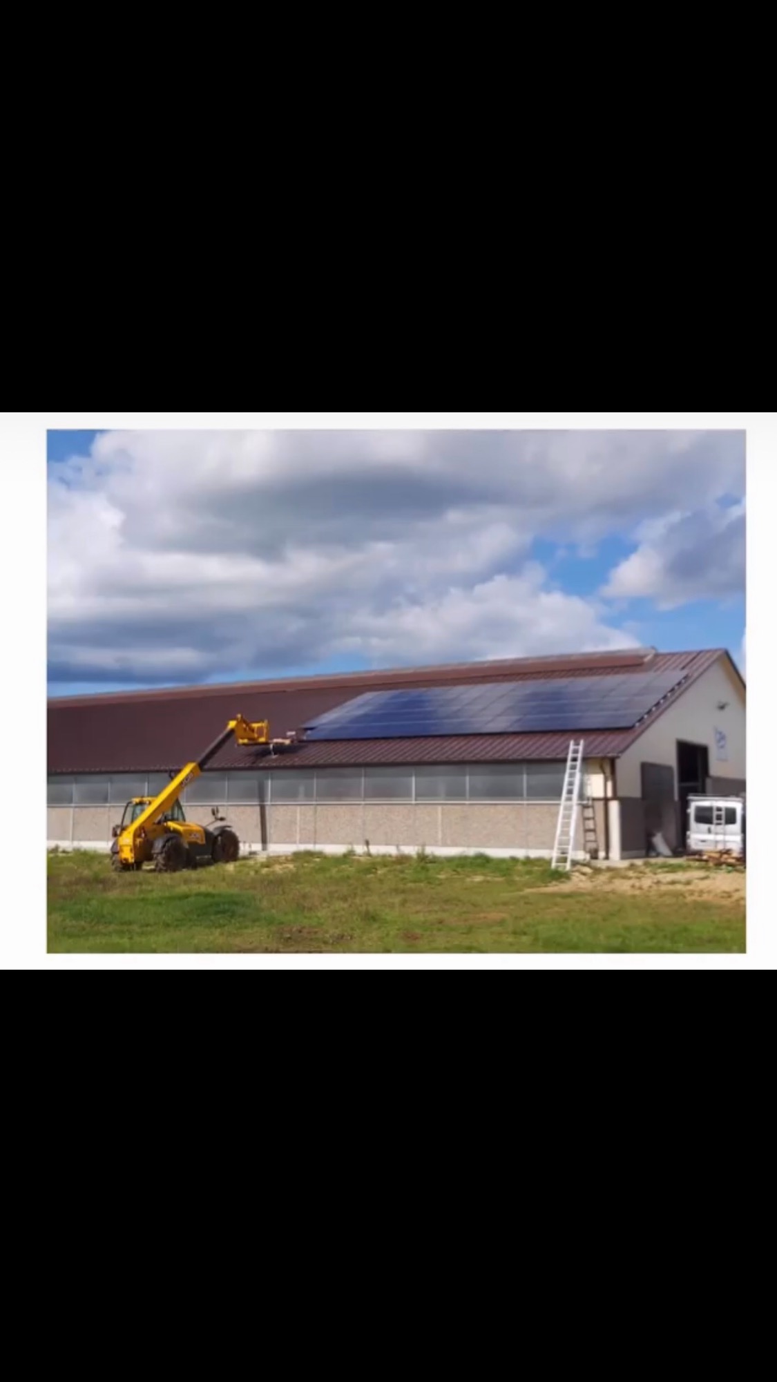 Żółty podnośnik teleskopowy z pracownikiem montującym panele fotowoltaiczne na dachu budynku gospodarczego, obok drabina oparta o ścianę, na tle pochmurnego nieba.