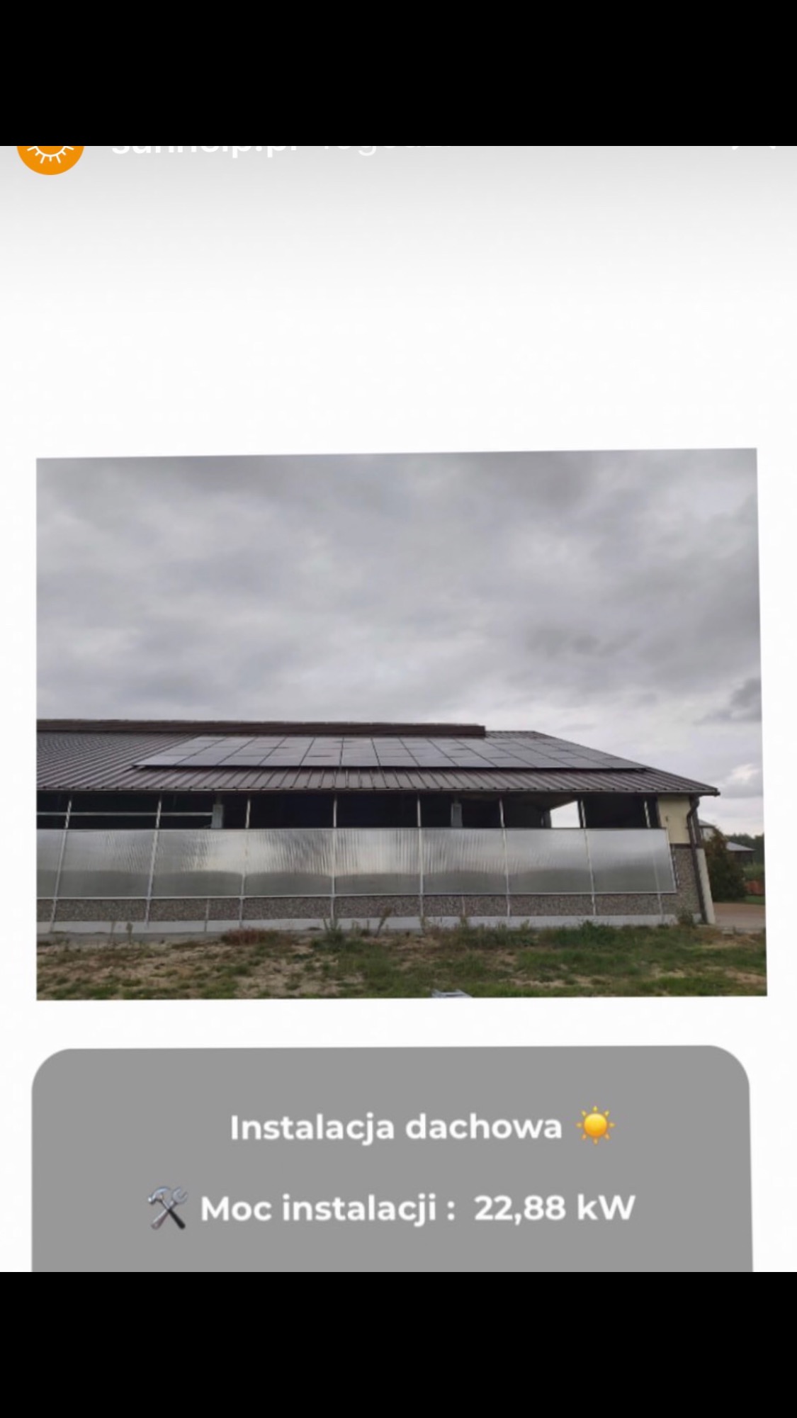 Dach budynku gospodarczego z brązowymi panelami słonecznymi, szara elewacja i pochmurne niebo w tle.