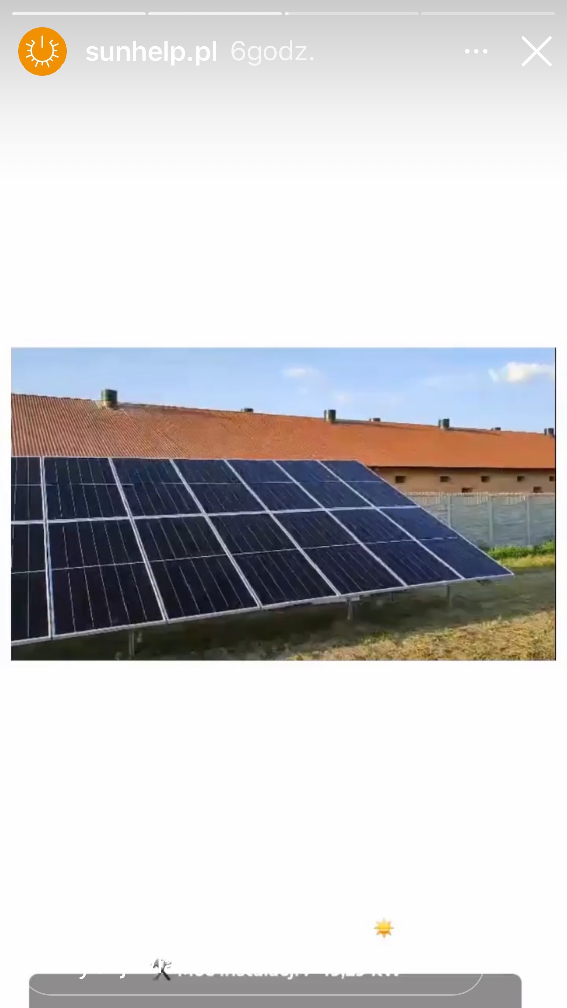 Rząd paneli słonecznych na metalowej konstrukcji, na tle budynku z czerwoną dachówką i betonowego ogrodzenia. Niebo jest jasne, bezchmurne.