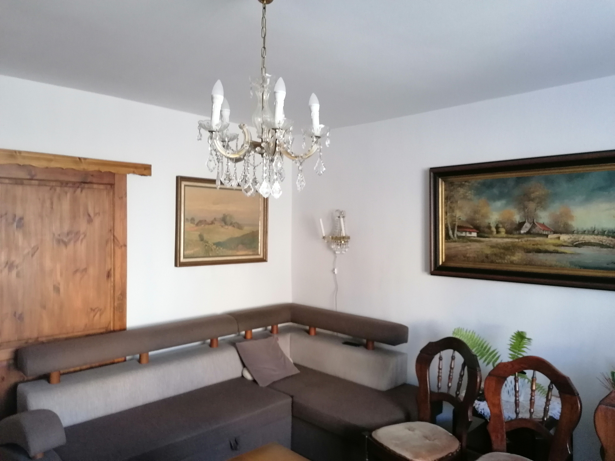 Jasne wnętrze pokoju z narożną sofą, drewnianymi krzesłami i obrazami na ścianach, oświetlone kryształowym żyrandolem.