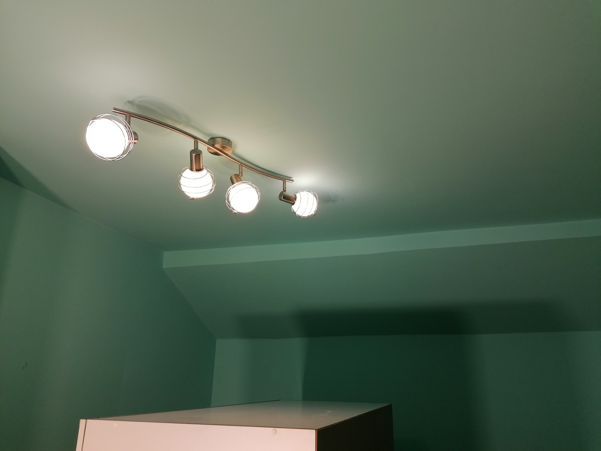 Świeżo pomalowane na zielono ściany i sufit poddasza, z widoczną lampą sufitową z czterema kulistymi reflektorami.
