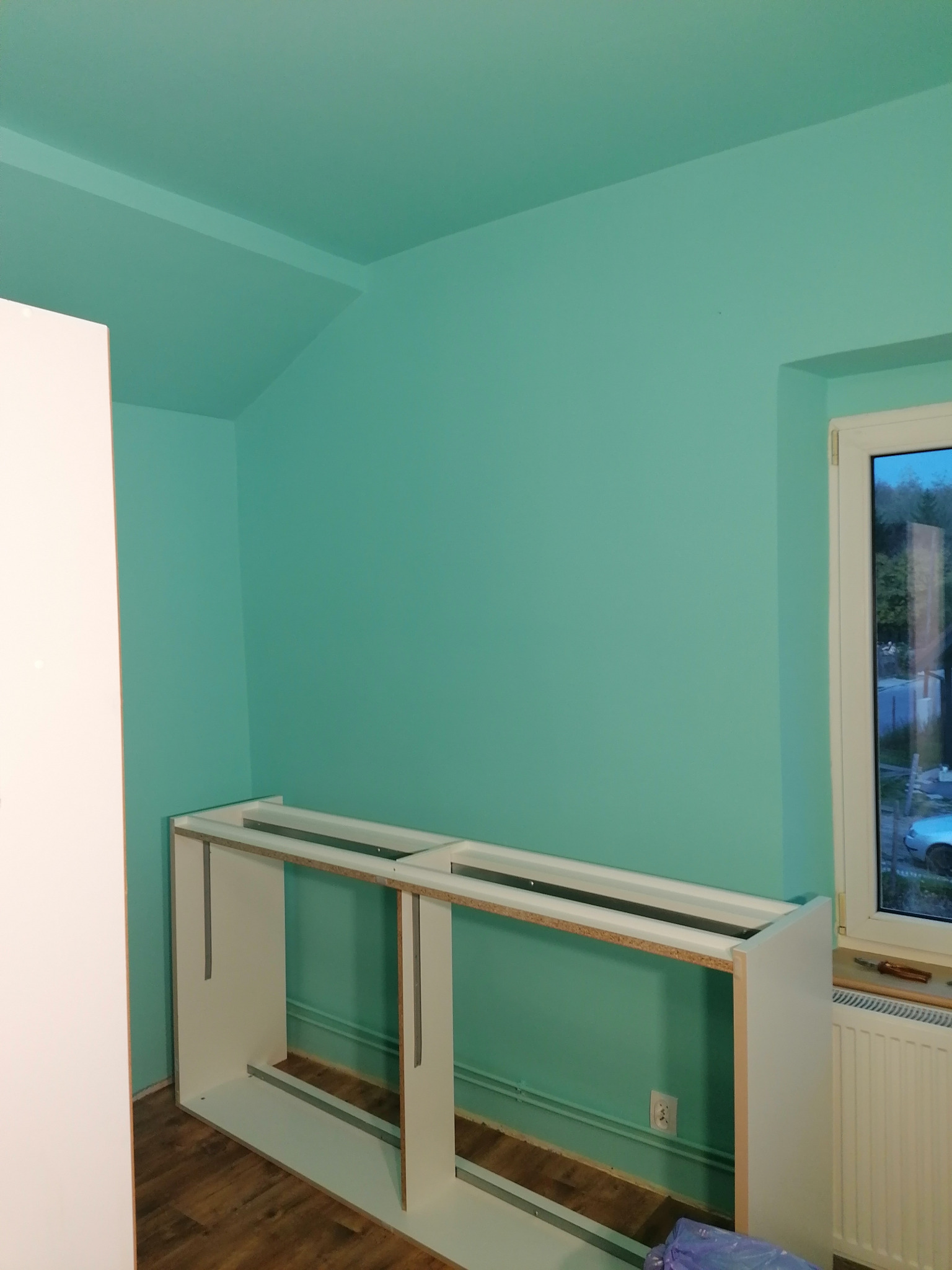 Świeżo pomalowane na turkusowo ściany pokoju z zamontowaną częścią białej zabudowy meblowej, widoczne gniazdko elektryczne i okno.