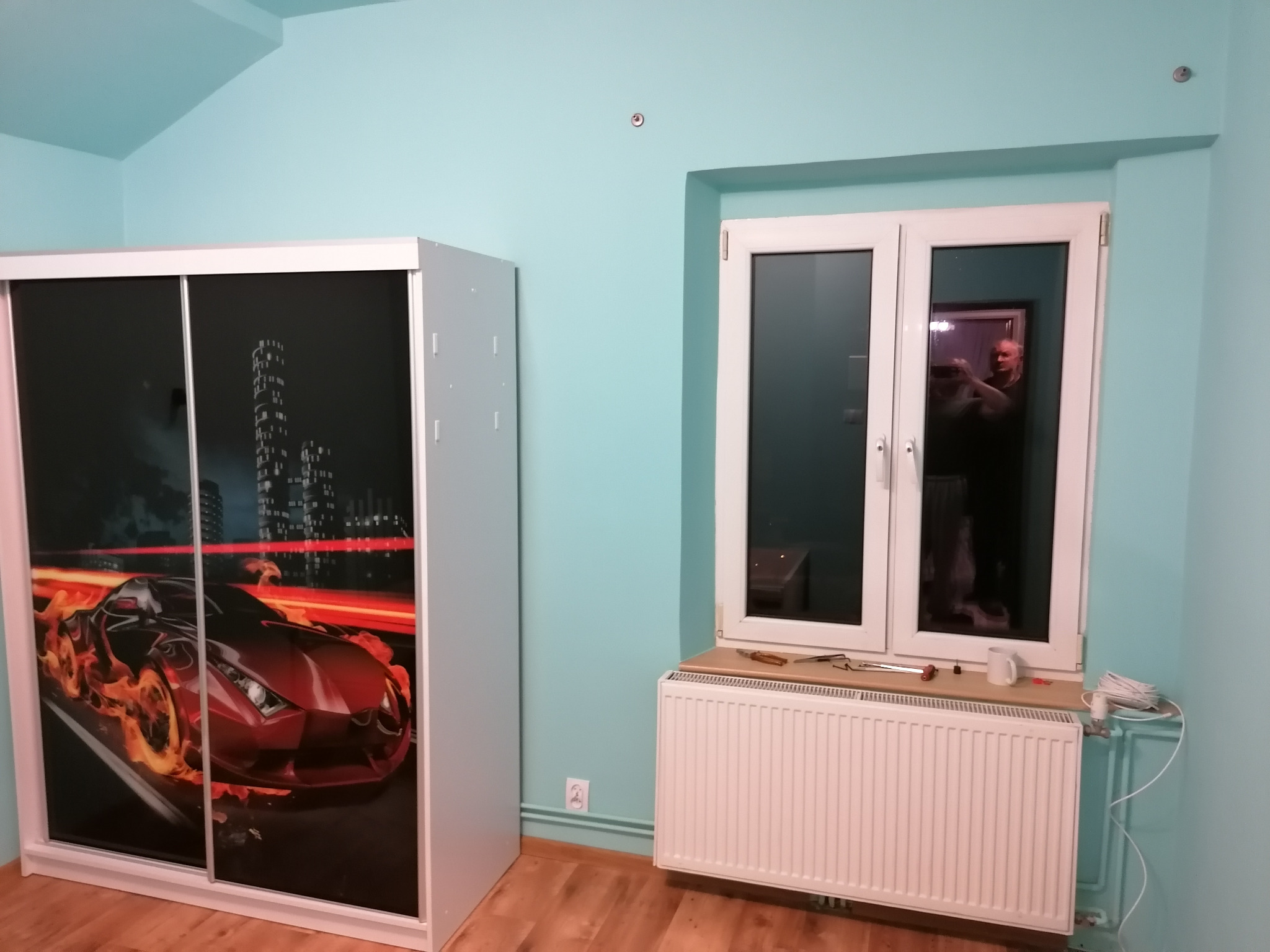 Pokój pomalowany na turkusowo z nowym oknem PCV, grzejnikiem, szafą przesuwną z grafiką płonącego samochodu i widocznym odbiciem fotografa w szybie okna.