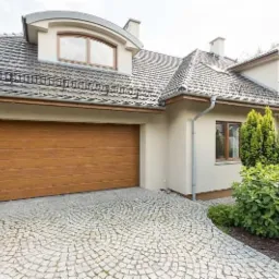 Nowoczesny dom jednorodzinny z brązową bramą garażową i podjazdem wyłożonym kostką granitową ułożoną w wachlarz, widoczne rynny i dachówka w kolorze grafitowym.