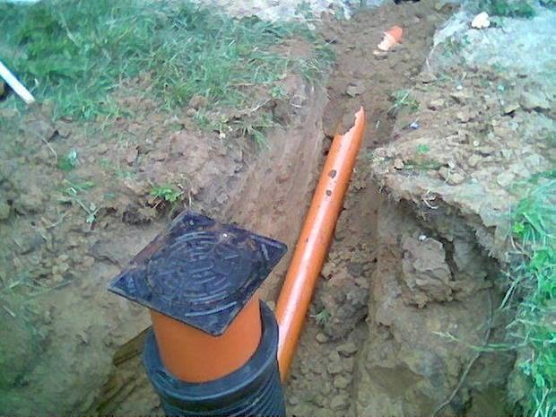 Pomarańczowa rura kanalizacyjna w wykopie z podłączoną czarną rurą karbowaną i studzienką rewizyjną z żeliwnym włazem, widok z góry na ziemię i trawę.