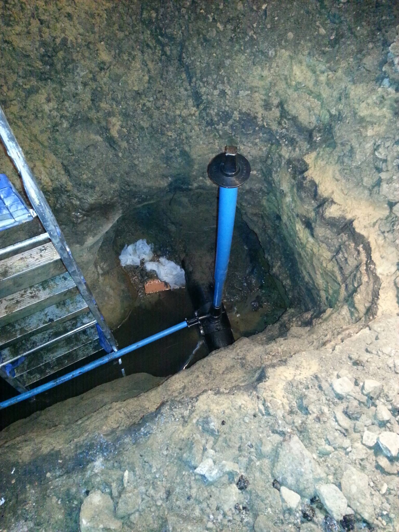 Wykop z drabinką, w którym widoczna jest niebieska rura pionowa z czarnym kołnierzem, podłączona do poziomej rury, w dole woda i gruz.