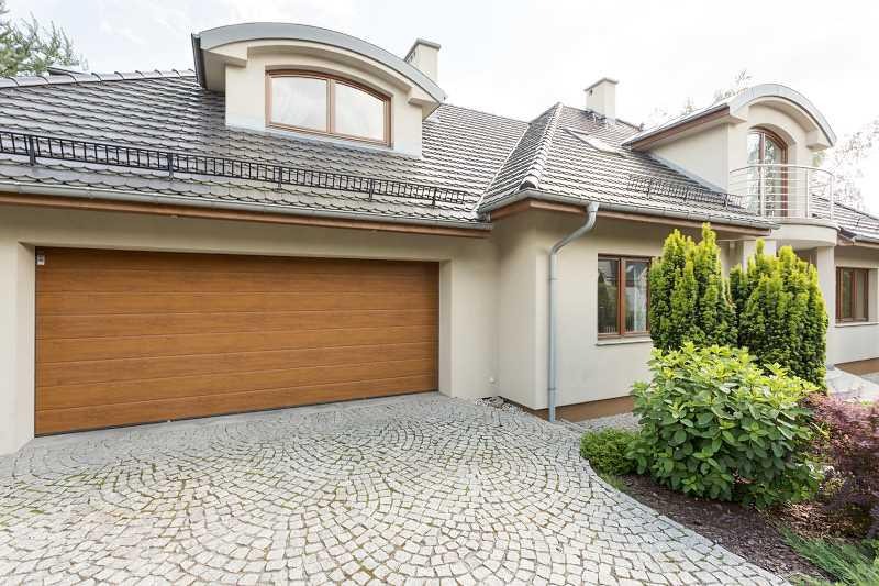 Nowoczesny dom jednorodzinny z brązową bramą garażową i podjazdem wyłożonym kostką granitową ułożoną w wachlarz, widoczne rynny i dachówka w kolorze grafitowym.
