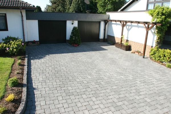Ułożona szara kostka brukowa na podjeździe przed domem z dwoma garażami, widoczna pergola z drewnianą konstrukcją po prawej stronie i zadbany trawnik z rabatami po lewej.