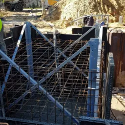 Wykop z szalunkiem i stalowym zbrojeniem klatkowym, częściowo zalany betonem, w tle betoniarka i hałda piachu.