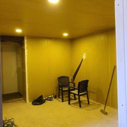 Michael - Pokój w trakcie prac wykończeniowych: ściany wyłożone płytami, widoczne narzędzia, dwie czarne krzesła ogrodowe w centrum, otwór w ścianie z wystającą łatą, oświetlenie sufitowe.