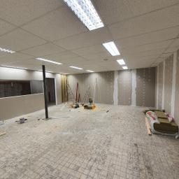 Michael - Wnętrze biura w trakcie remontu: widoczne płyty kartonowo-gipsowe na ścianach, podwieszany sufit kasetonowy z oświetleniem rastrowym, narzędzia budowlane i materiały izolacyjne na podłodze...