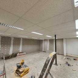 Michael - Wnętrze remontowanego biura, widoczne ściany z częściowo położoną gładzią, sufit kasetonowy z oświetleniem, narzędzia budowlane i drabiny na podłodze.