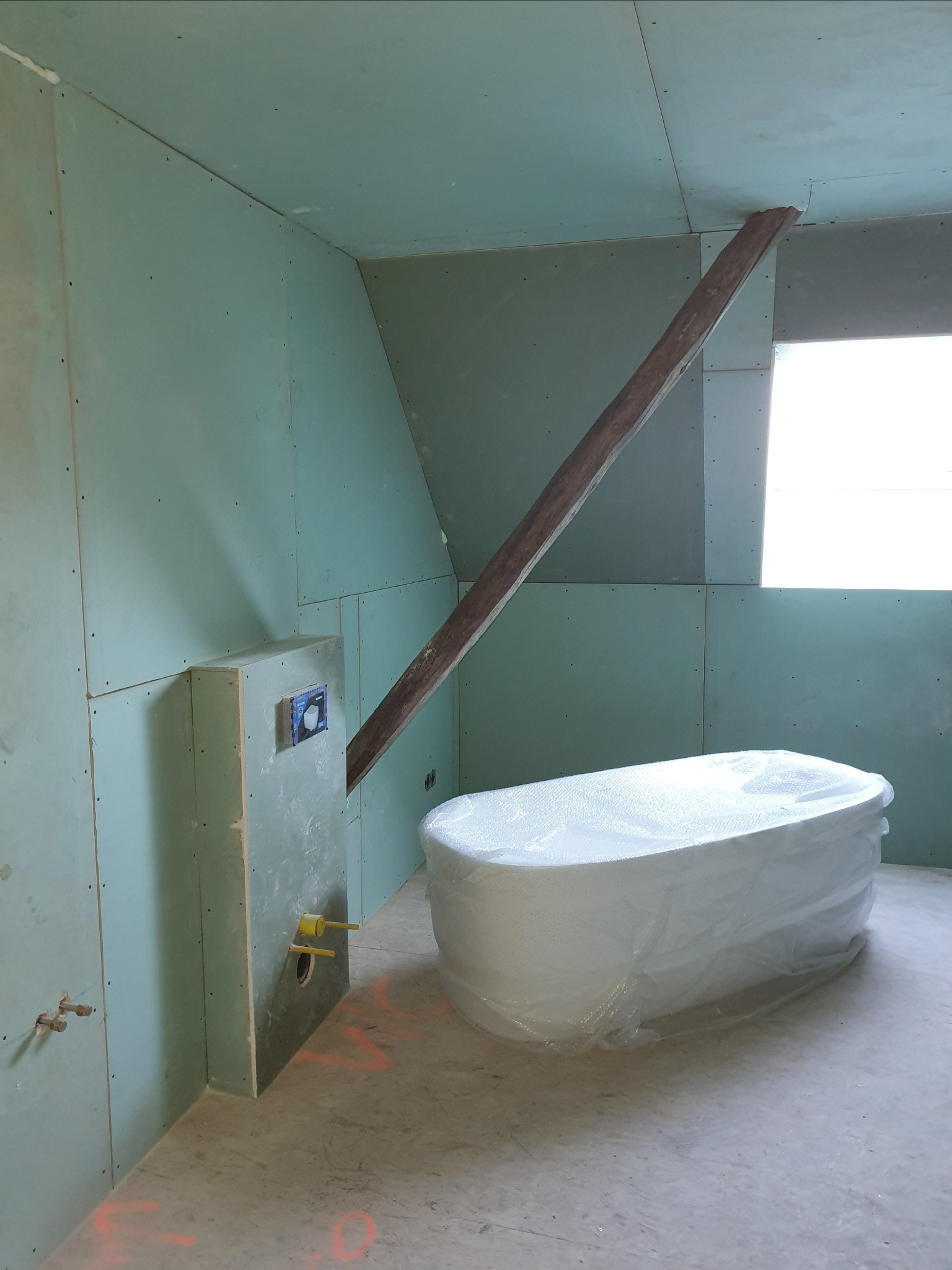 Biała wanna owinięta folią ochronną w surowej łazience z płyt gipsowo-kartonowych, drewniana belka podtrzymująca sufit, widoczne elementy instalacji wodnej.