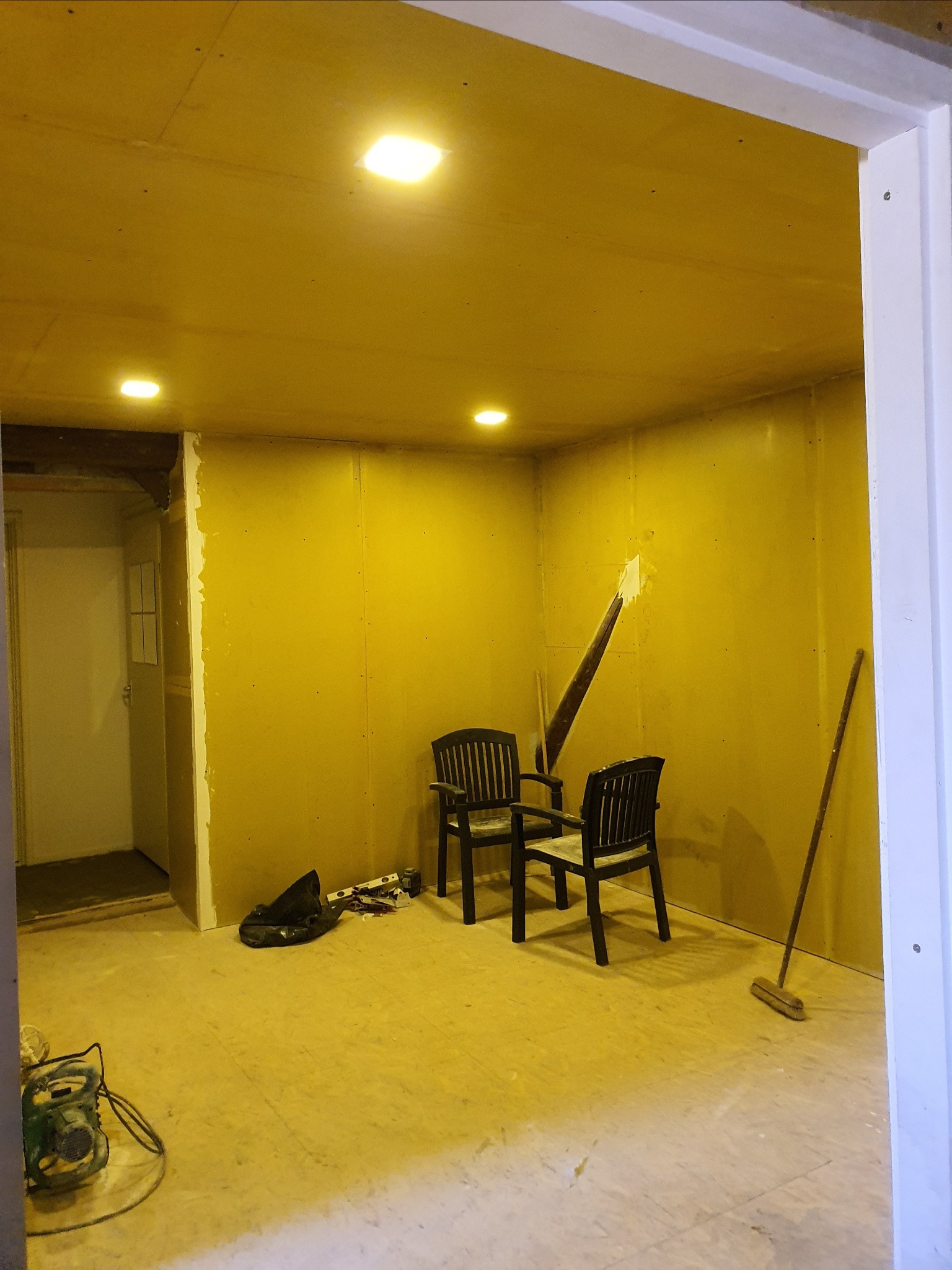 Pokój w trakcie prac wykończeniowych: ściany wyłożone płytami, widoczne narzędzia, dwie czarne krzesła ogrodowe w centrum, otwór w ścianie z wystającą łatą, oświetlenie sufitowe.