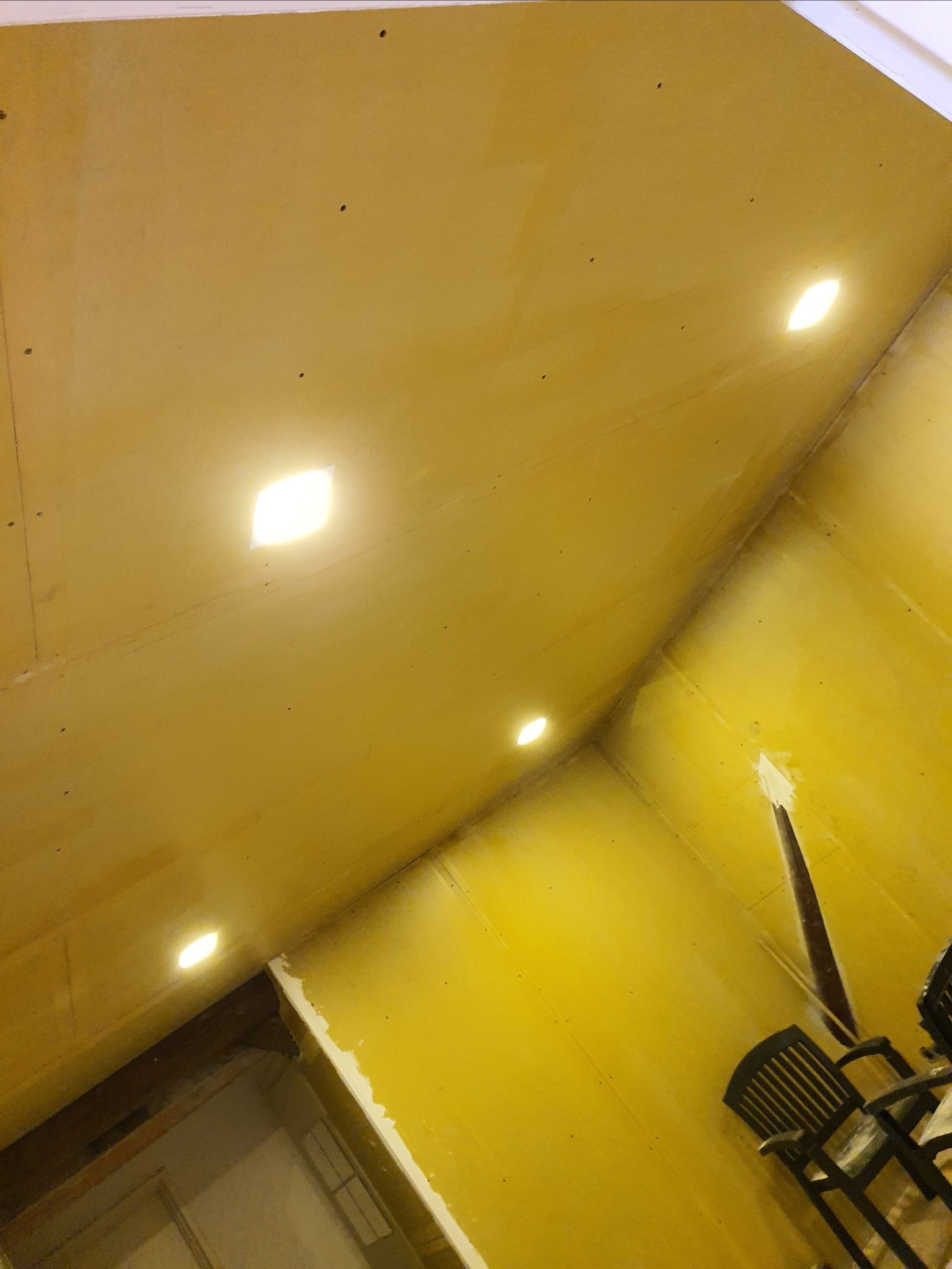 Wnętrze w trakcie prac wykończeniowych; ściany i sufit pokryte gładzią szpachlową, widoczne otwory pod oświetlenie punktowe w suficie, krzesło w rogu pomieszczenia.