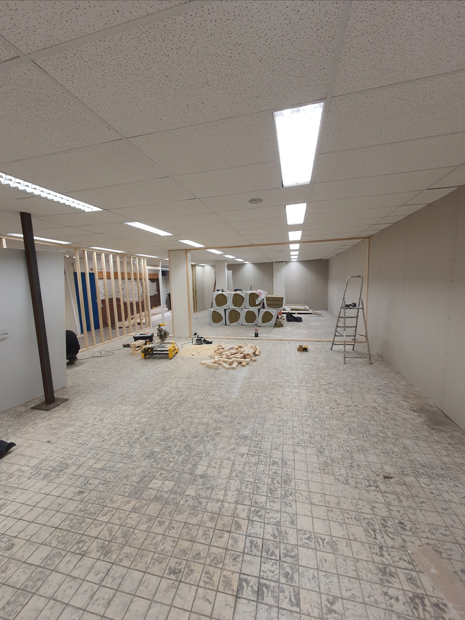 Przestrzeń biurowa w trakcie remontu: widoczne elementy konstrukcyjne ścian działowych, materiały izolacyjne w rolkach, narzędzia budowlane i drabina rozstawiona w pustym pomieszczeniu...