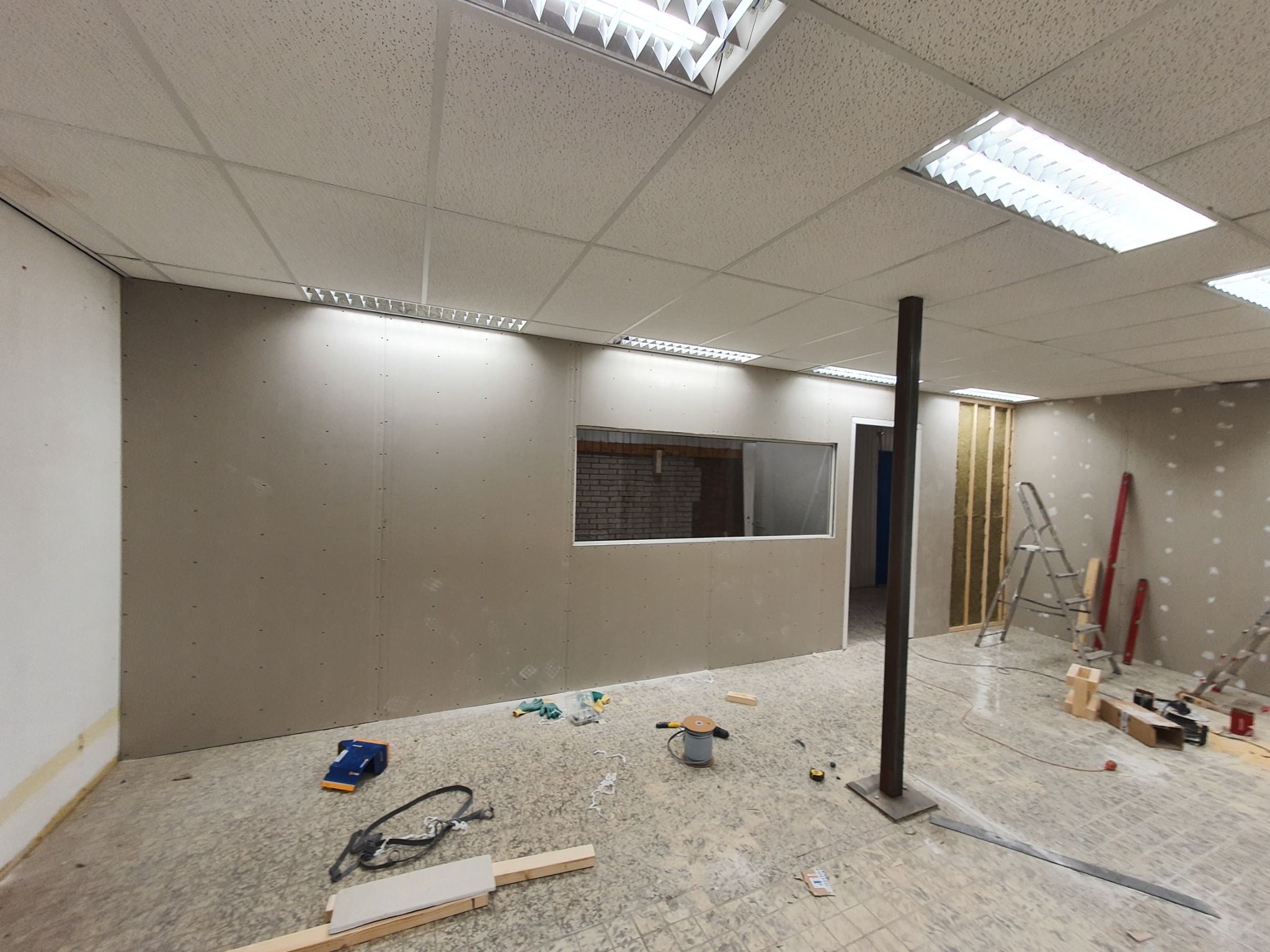 Wnętrze biura w trakcie remontu: sufit podwieszany kasetonowy, ściany z płyt gipsowo-kartonowych z oknem, widoczne narzędzia i materiały budowlane, podłoga częściowo zabezpieczona.
