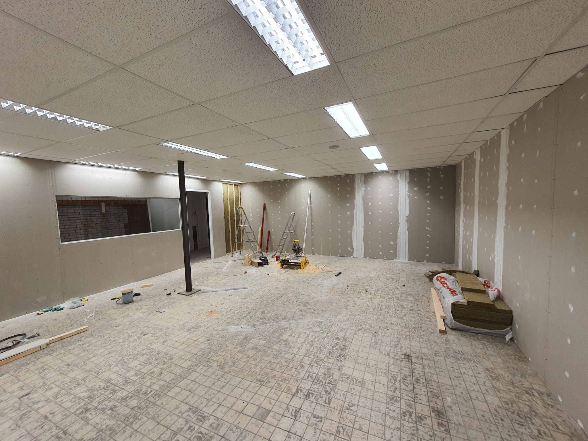 Wnętrze biura w trakcie remontu: widoczne płyty kartonowo-gipsowe na ścianach, podwieszany sufit kasetonowy z oświetleniem rastrowym, narzędzia budowlane i materiały izolacyjne na podłodze...