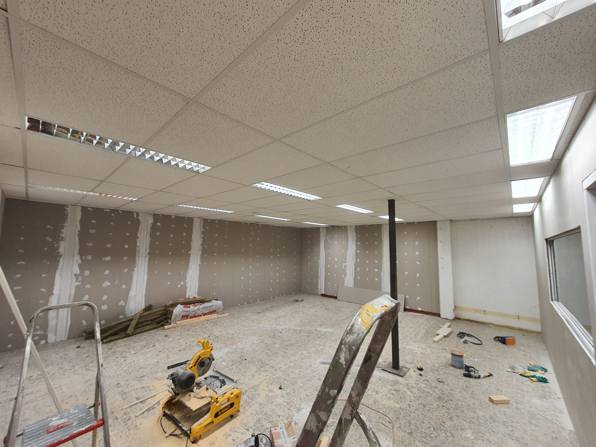 Wnętrze remontowanego biura, widoczne ściany z częściowo położoną gładzią, sufit kasetonowy z oświetleniem, narzędzia budowlane i drabiny na podłodze.