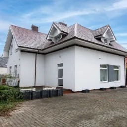 Nowoczesny, biały budynek z brązowym dachem, widok z zewnątrz na tle błękitnego nieba z chmurami, kostka brukowa na podjeździe.