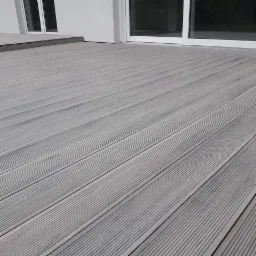 Szara deska tarasowa z ryflowaną powierzchnią, ułożona równolegle do białych, przesuwnych drzwi tarasowych i okna, tworząca nowoczesną przestrzeń zewnętrzną.