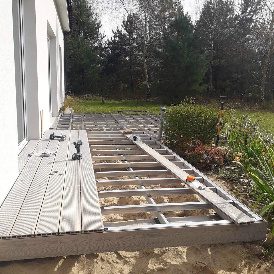 Montaż kompozytowego tarasu zewnętrznego: widoczne elementy konstrukcyjne, częściowo ułożone deski, wkrętarka i poziomica na tle ogrodu z drzewami.