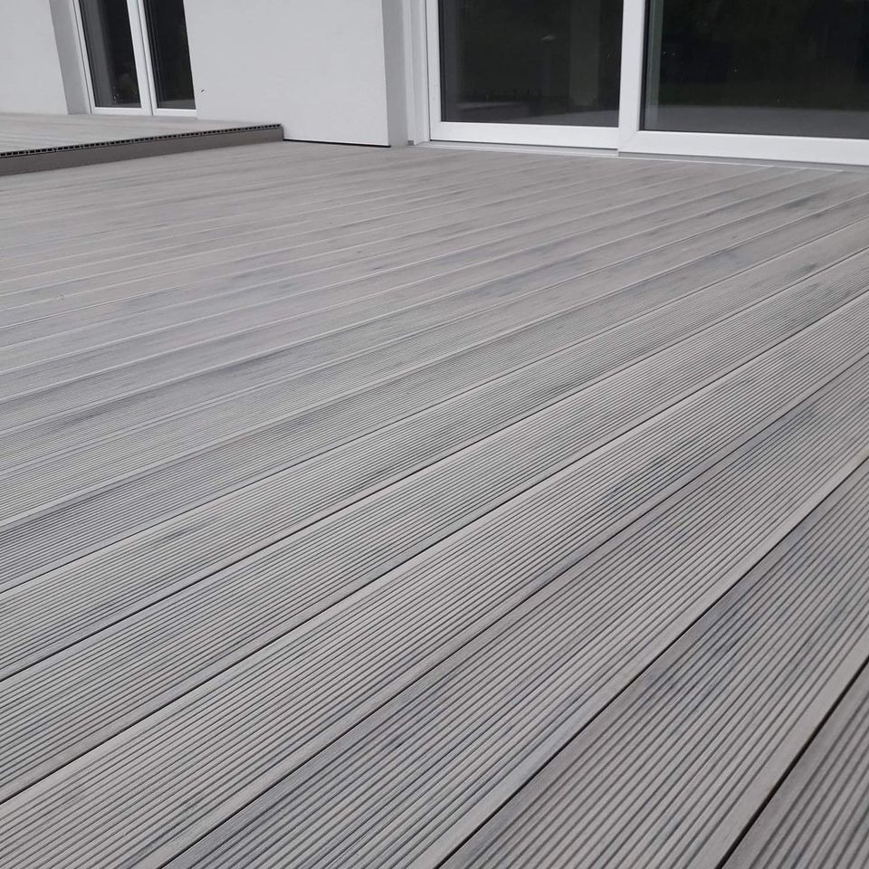 Szara deska tarasowa z ryflowaną powierzchnią, ułożona równolegle do białych, przesuwnych drzwi tarasowych i okna, tworząca nowoczesną przestrzeń zewnętrzną.