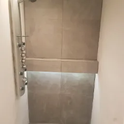 Nowoczesny prysznic z szarą, kwadratową glazurą, chromowaną kolumną natryskową z hydromasażem i półką podświetloną od dołu.