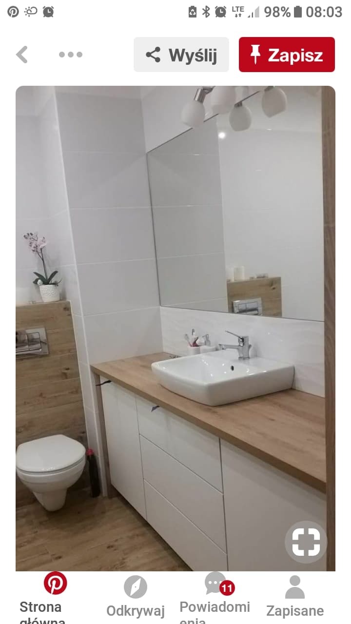 Nowoczesna łazienka z białymi płytkami, drewnopodobną zabudową toalety i blatem, oraz kwadratową umywalką nablatową i dużym lustrem.