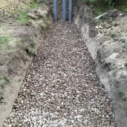 Wykop wypełniony żwirem z wystającymi pionowo trzema szarymi rurami, widoczne ślady ziemi po bokach wykopu i trawa w tle.