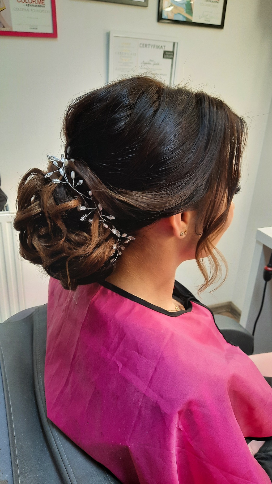 Upięcie ślubne z ciemnych włosów, ozdobione srebrną gałązką z kryształkami, widok z tyłu, klientka w różowym okryciu ochronnym.