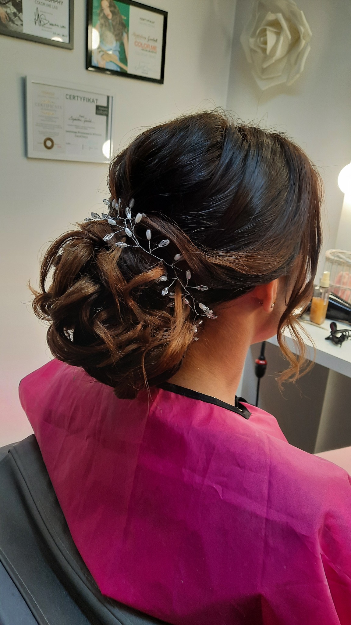 Upięcie włosów w kok z ozdobną spinką z kryształkami, widok z tyłu, różowa peleryna ochronna na ramionach klientki.