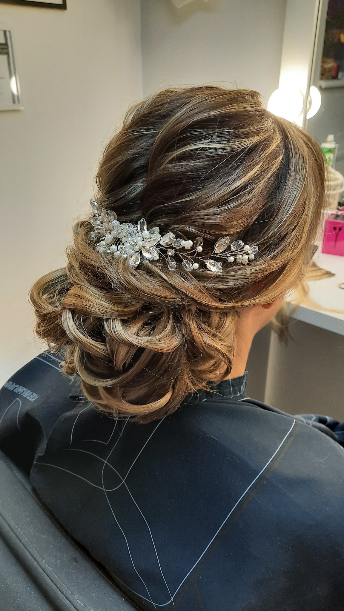 Upięcie ślubne z brązowych włosów z blond pasemkami, ozdobione delikatną spinką z kryształkami i perłami, widok z tyłu, klientka w czarnej pelerynie fryzjerskiej.