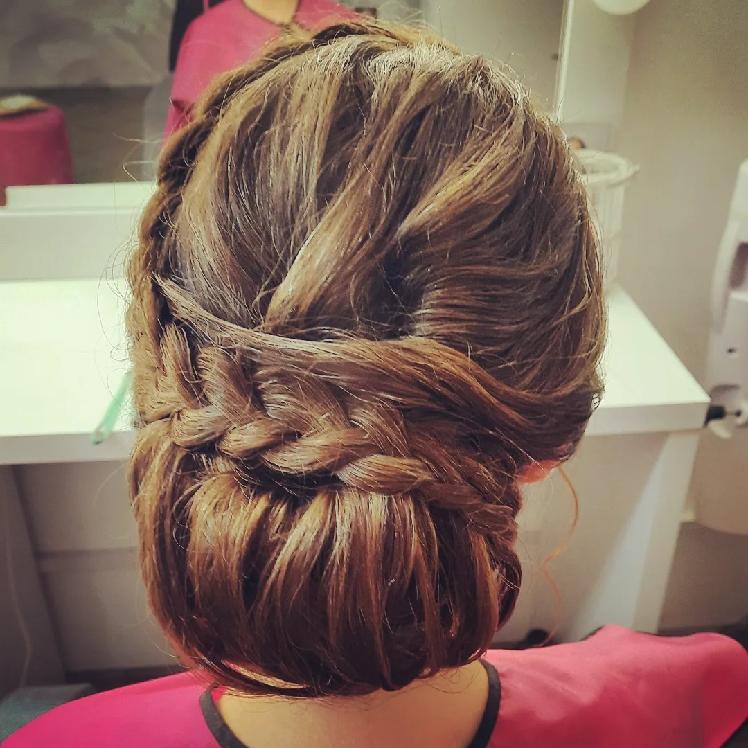 Upięcie włosów z warkoczem na brązowych włosach, widok z tyłu, różowa peleryna ochronna na ramionach.