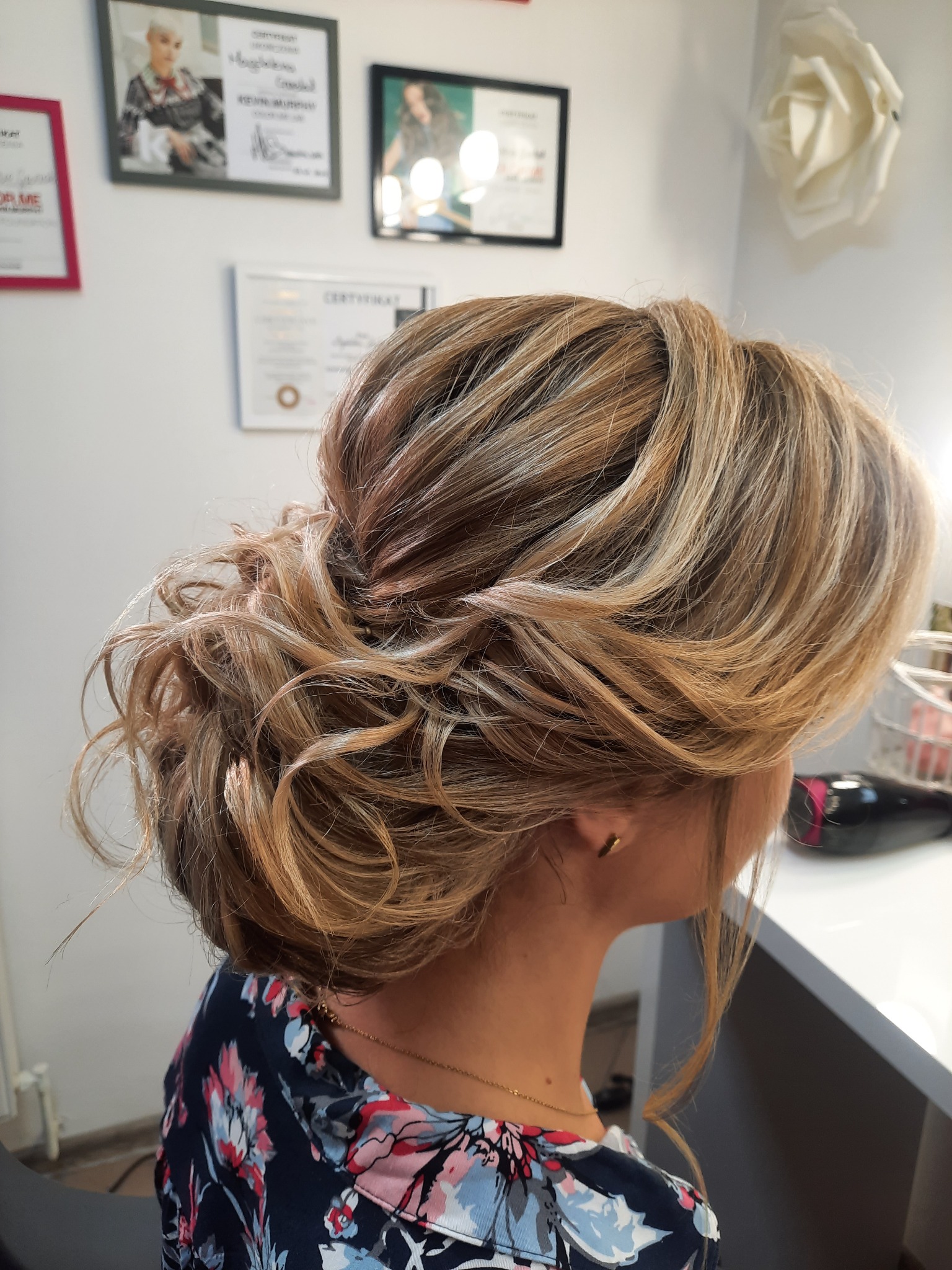 Upięcie włosów blond z pasemkami, widok z boku, delikatne loki i upięcie w kok, w tle certyfikaty i zdjęcia na ścianie.