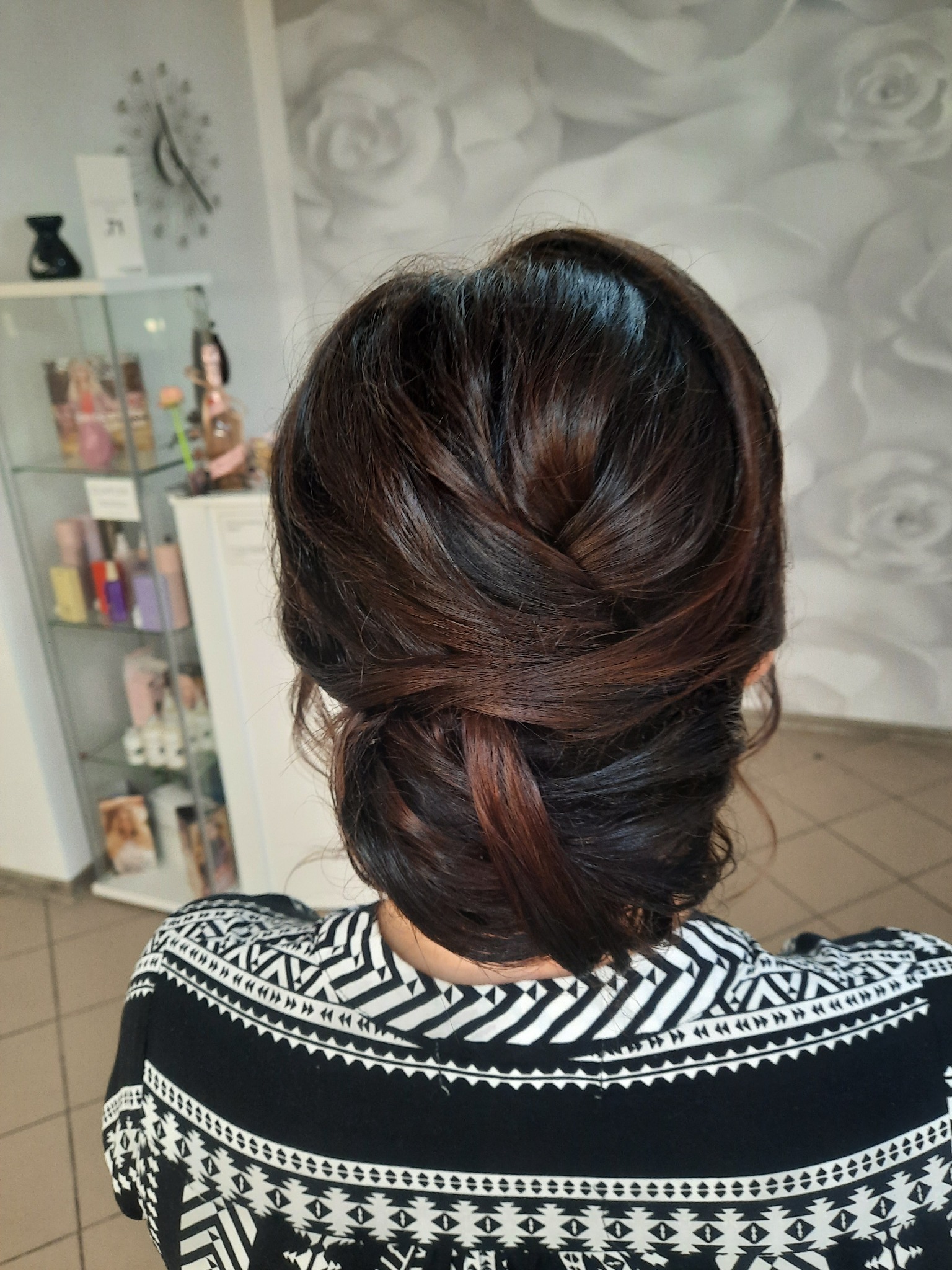 Upięcie włosów w kok z warkoczem, widok z tyłu, na tle ściany w kwiaty i przeszklonej szafki z kosmetykami.