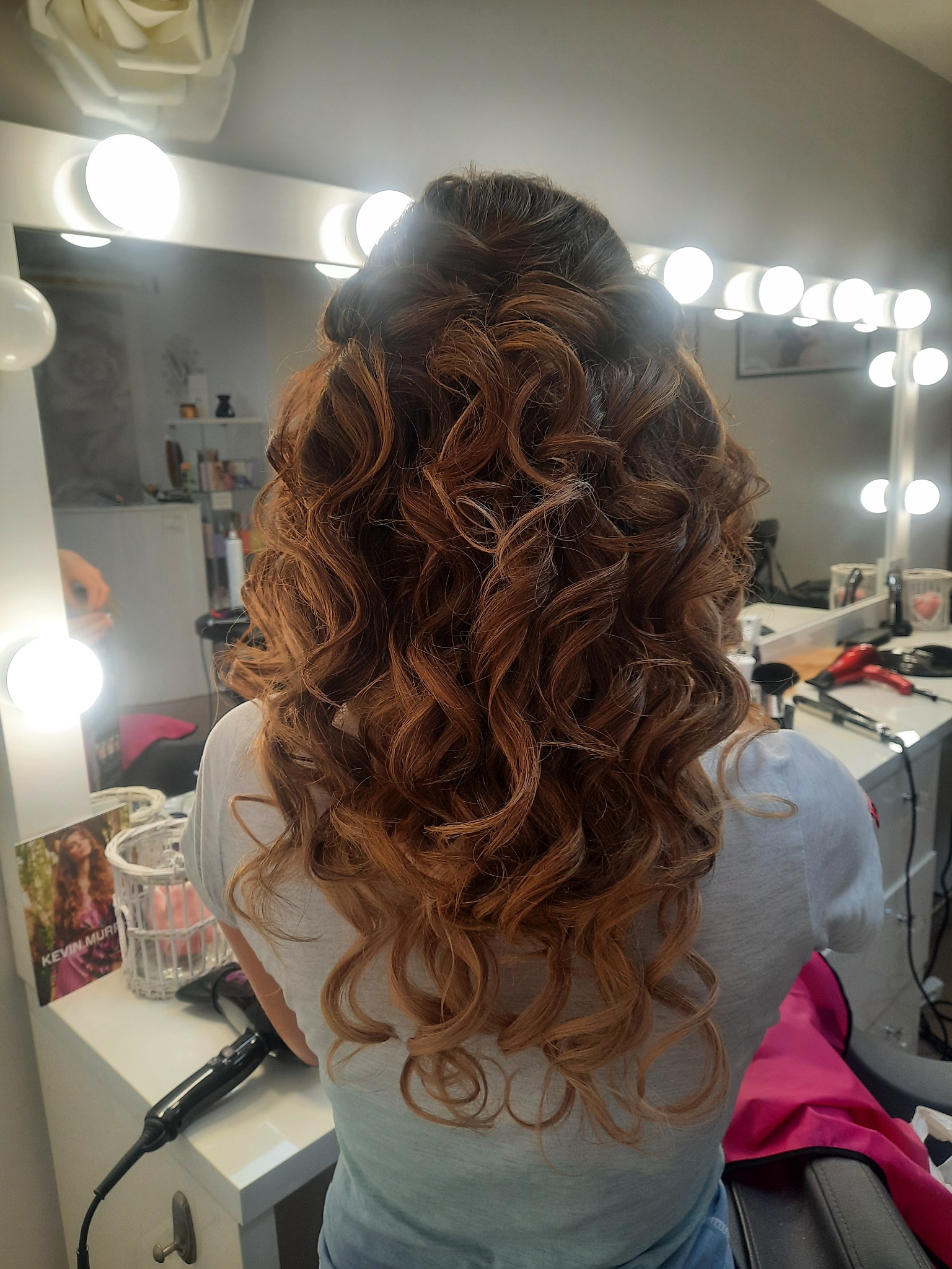 Kobieca fryzura z loków w odcieniach brązu i blondu, upięta z tyłu głowy, widoczna na tle lustra z oświetleniem w salonie.