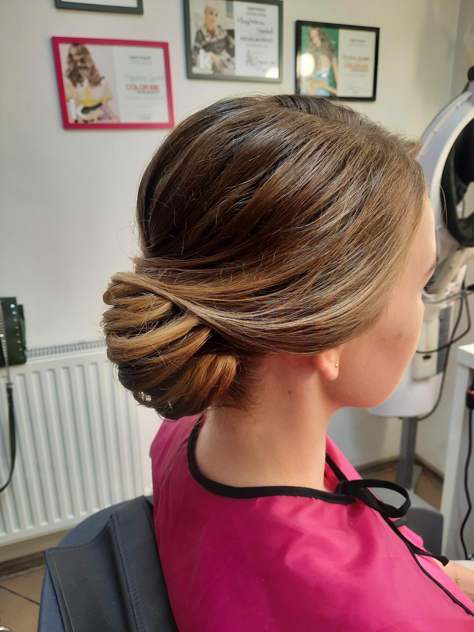 Upięcie włosów w kok z boku głowy, widoczne pasemka w odcieniach blondu, różowa peleryna ochronna, w tle ściana z ramkami i sprzęt fryzjerski.
