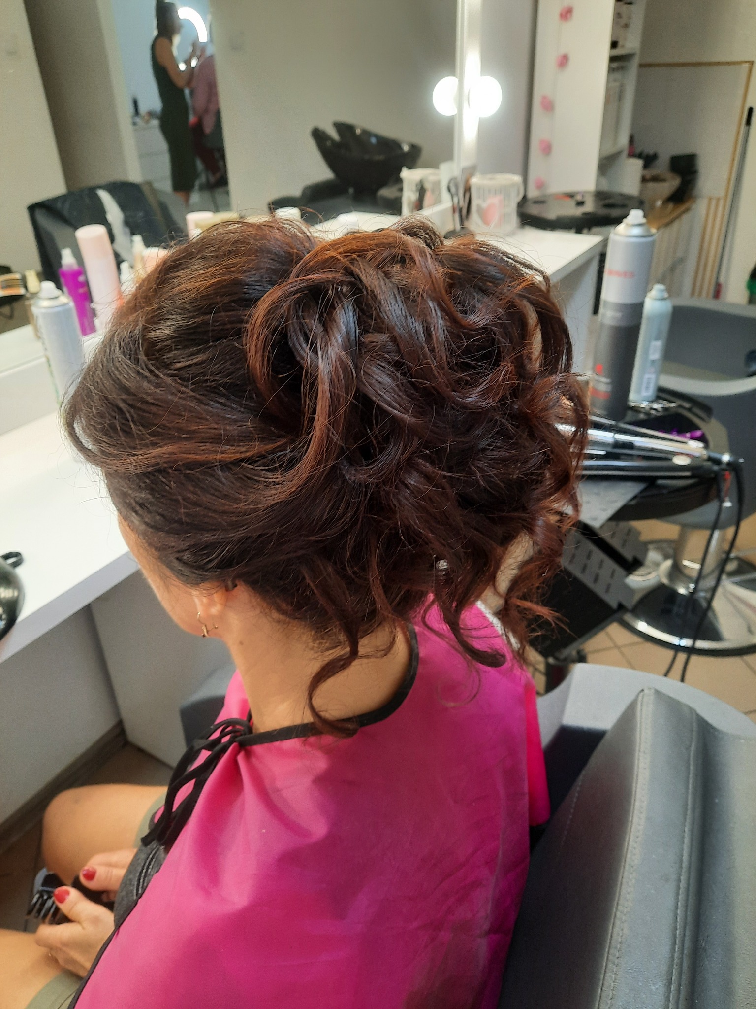 Upięcie włosów w odcieniach brązu z refleksami, widoczne z tyłu głowy, klientka w różowym okryciu ochronnym w salonie, w tle lustro i oświetlenie.