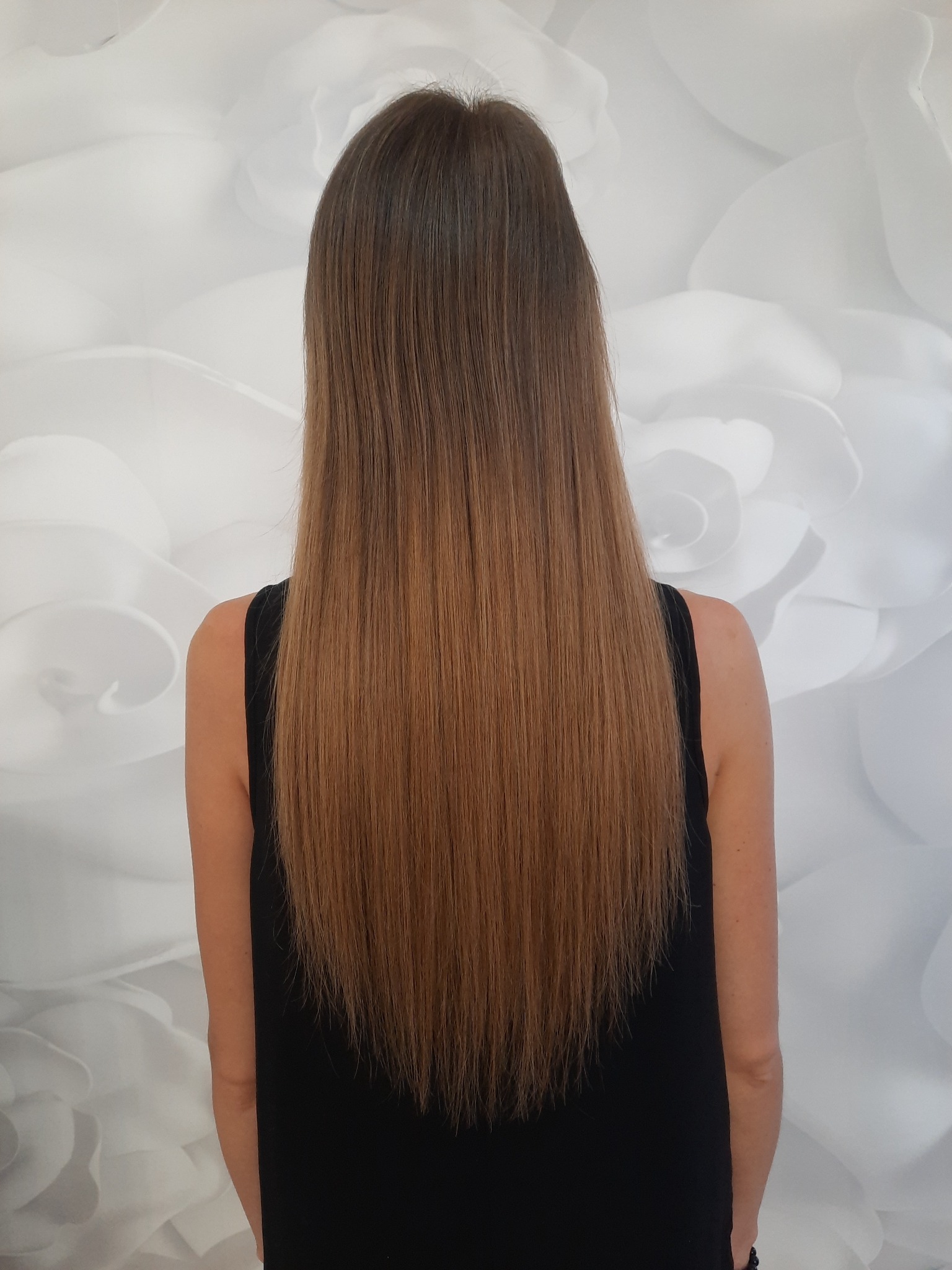 Długie, proste włosy w odcieniach brązu, widok z tyłu na tle białej ściany z motywem kwiatowym.
