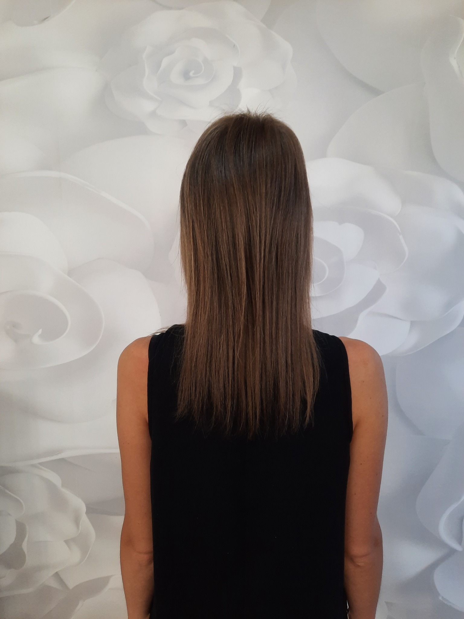 Widok z tyłu na proste, brązowe włosy kobiety, świeżo po strzyżeniu, na tle ściany z białych, papierowych róż.