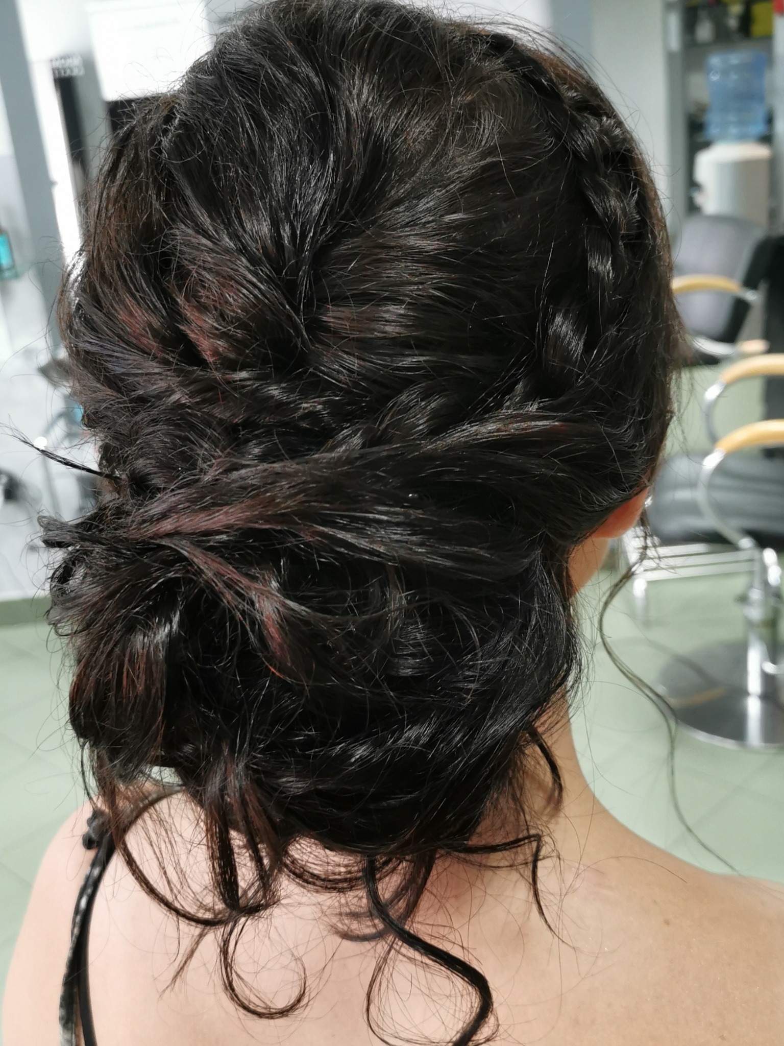 Upięcie z ciemnych włosów z widocznymi refleksami w odcieniach czerwieni, zebrane w kok z warkoczem, widok z tyłu na kark i ramiona klientki w salonie.