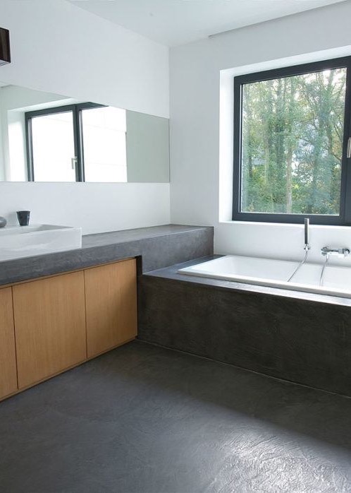Minimalistyczna łazienka z betonową posadzką, drewnianą szafką pod umywalką i wanną przy oknie z widokiem na drzewa.