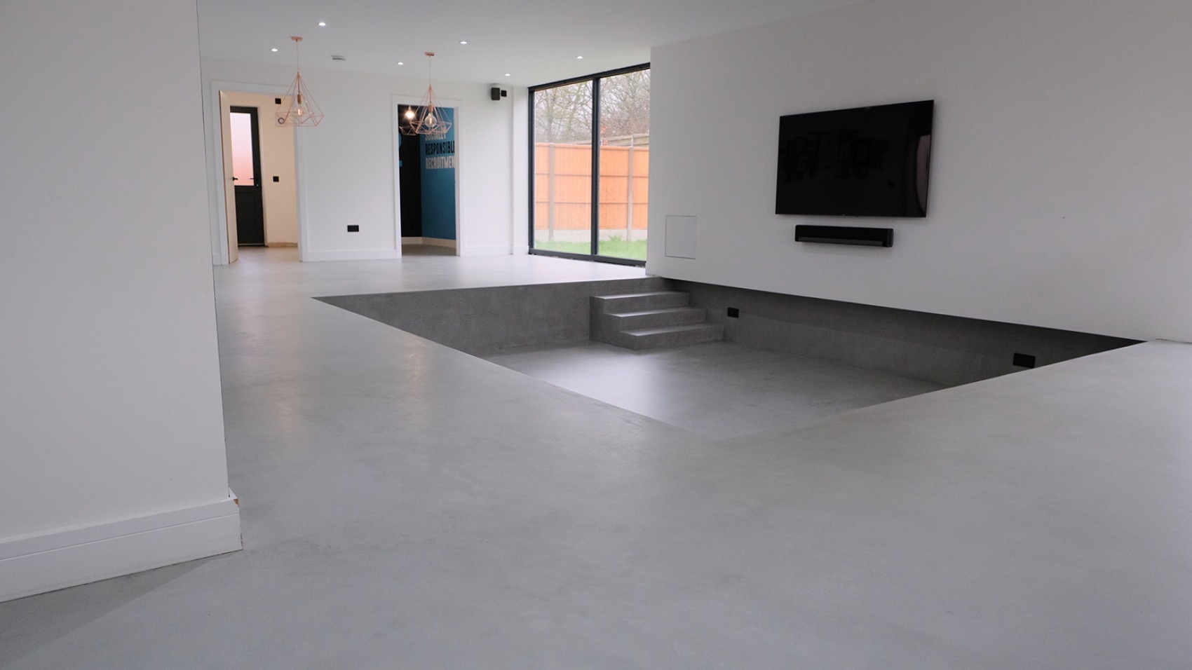 Nowoczesne wnętrze z szarą posadzką żywiczną i obniżonym poziomem podłogi z betonowymi schodami, minimalistyczny design z białymi ścianami i dużym oknem.