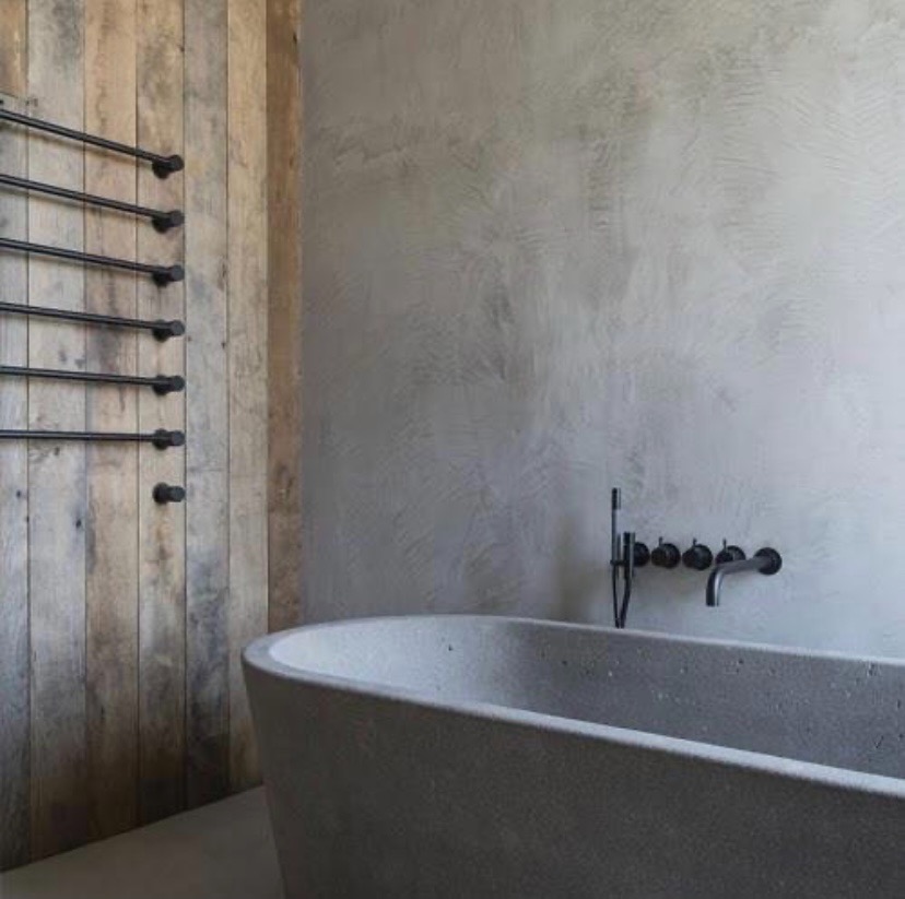 Minimalistyczna łazienka z betonową wanną wolnostojącą, ciemną armaturą ścienną i drewnianą ścianą z czarnym grzejnikiem drabinkowym na ręczniki.