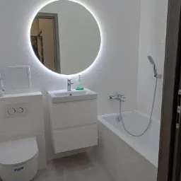 Nowoczesna, biała łazienka z podświetlanym okrągłym lustrem, wiszącą toaletą, umywalką z szafką i wanną z prysznicem; na ścianie widoczna paczka elastycznej fugi cementowej.