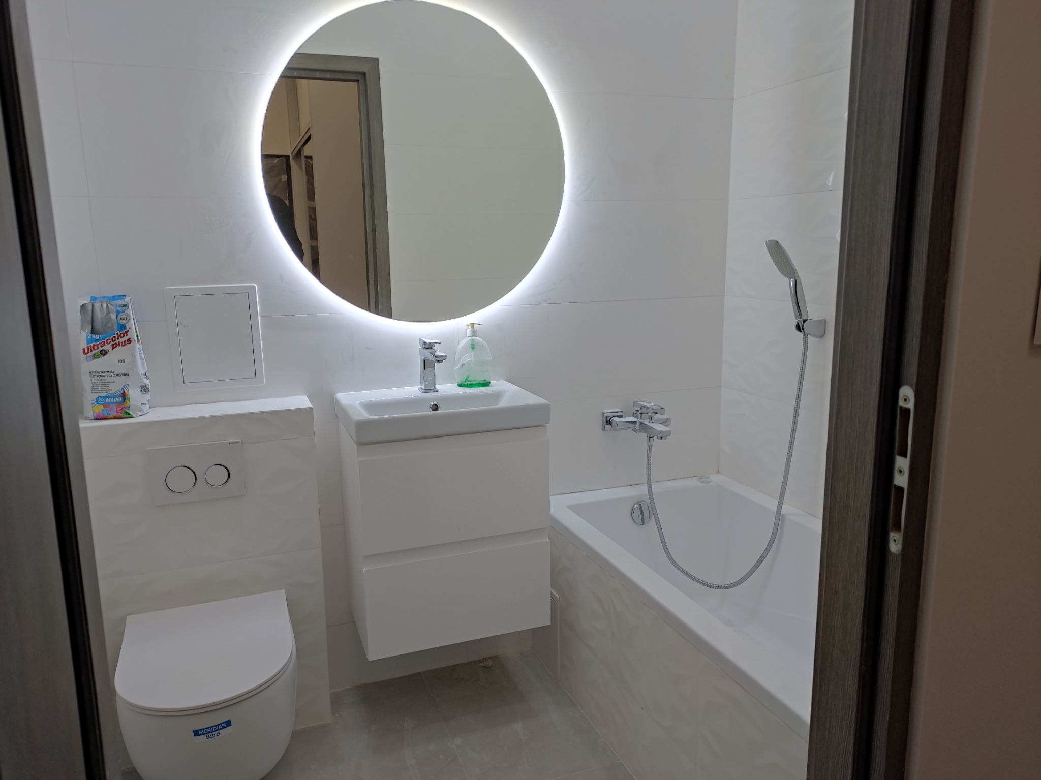 Nowoczesna, biała łazienka z podświetlanym okrągłym lustrem, wiszącą toaletą, umywalką z szafką i wanną z prysznicem; na ścianie widoczna paczka elastycznej fugi cementowej.