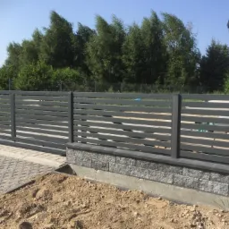 Nowoczesna, pozioma brama przesuwna z metalowymi lamelami w kolorze grafitowym, zamontowana na murku z bloczków betonowych, z widocznym podjazdem z kostki brukowej i nasypanym piaskiem.
